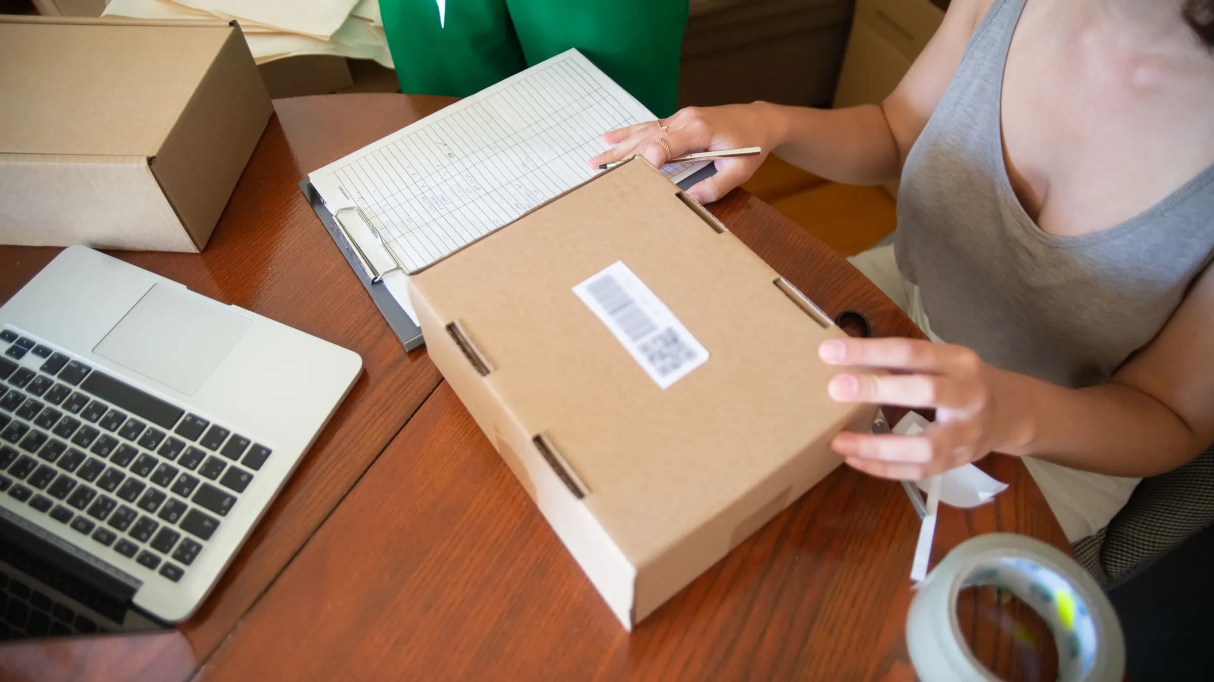 Hands pack an order into a cardboard box next to a laptop, an order sheet on a clipboard and a roll of tape.
