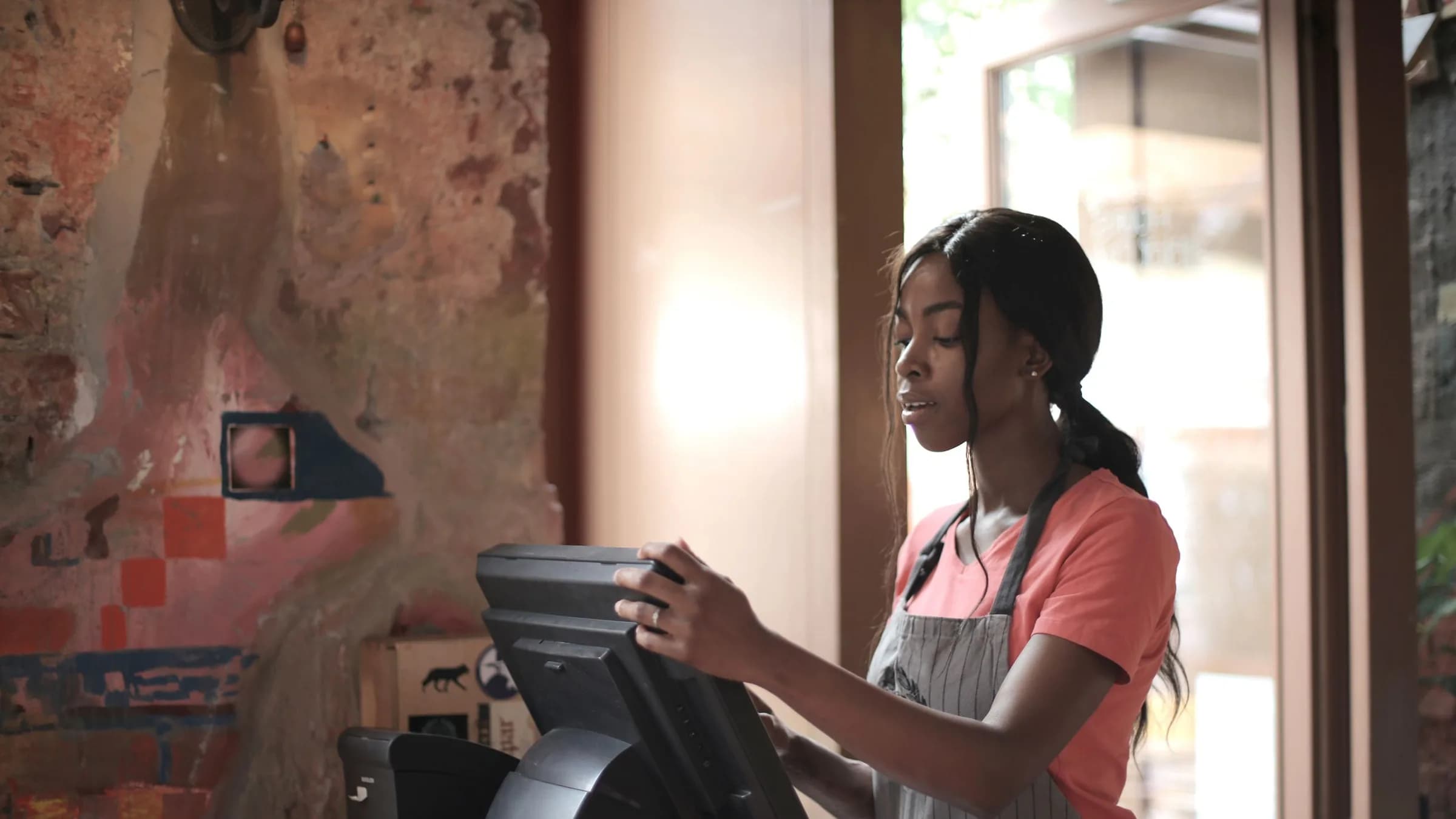 A cashier in an apron works the till screen at the counter of a small cafe.