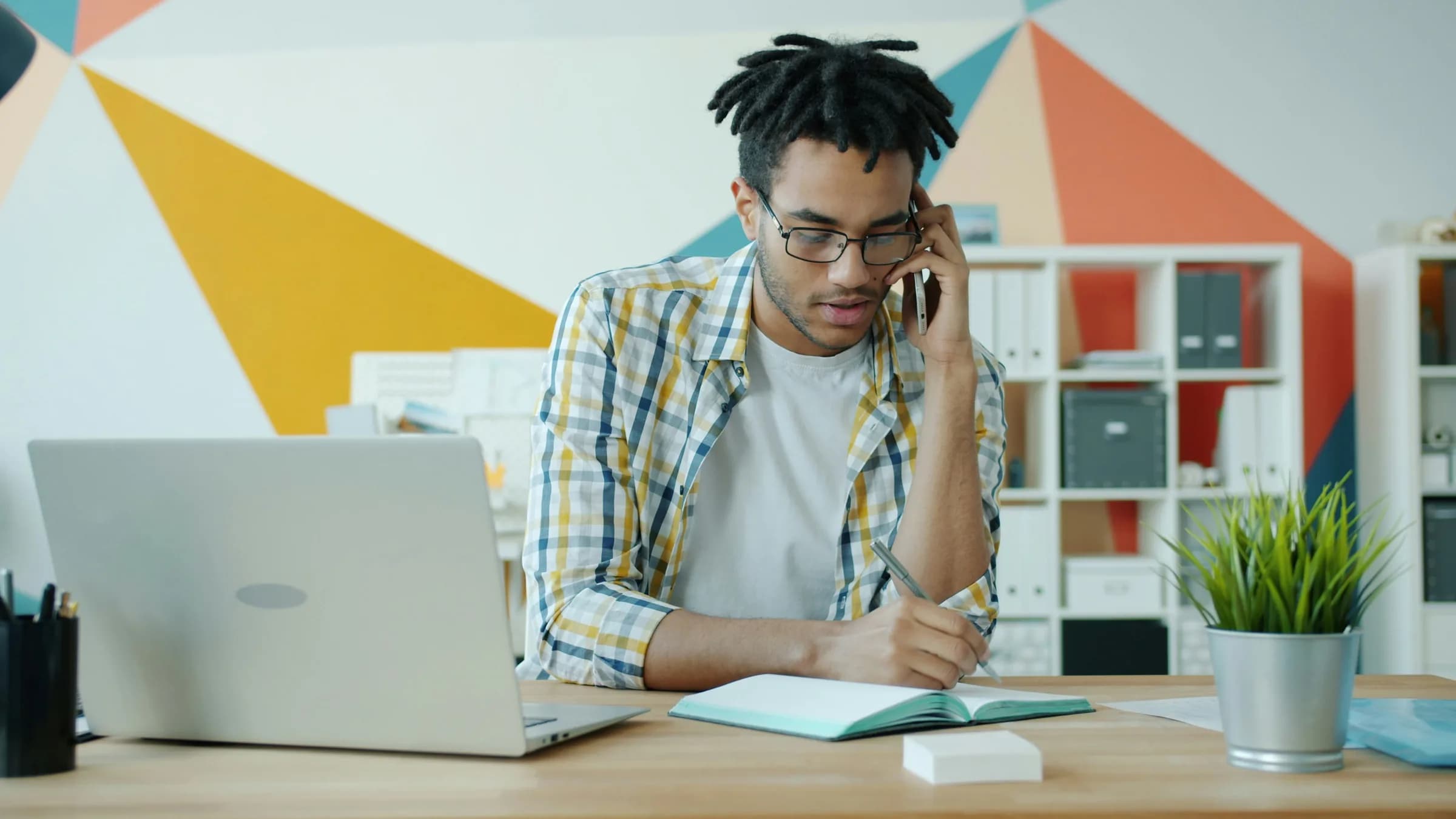 A business owner on the phone takes notes at a desk beside an open laptop.