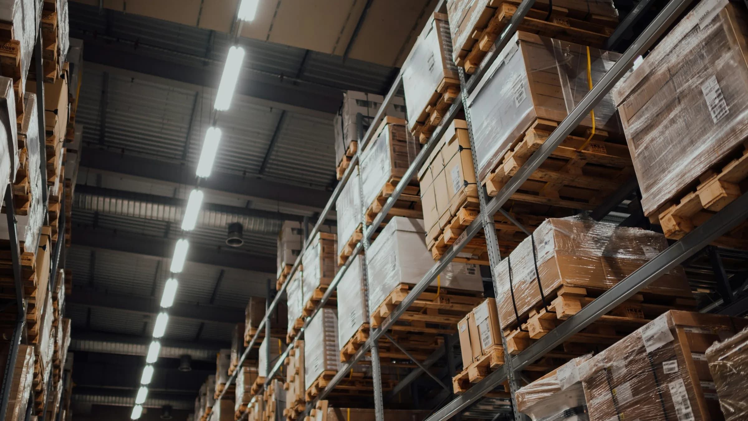 Tall warehouse racking stacked with wrapped cartons on wooden pallets under a line of ceiling lights.