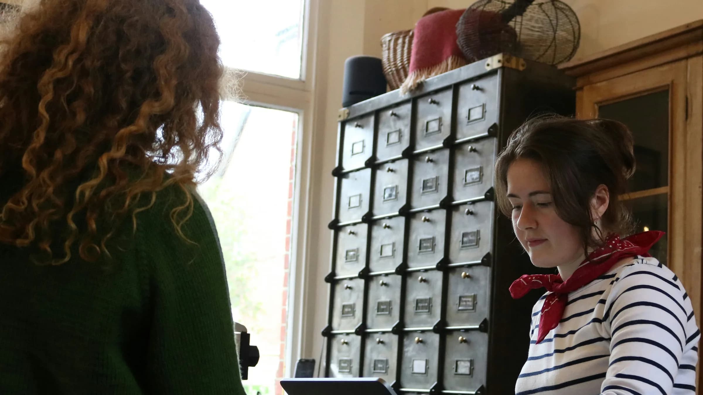 A shop assistant in a striped top serves a customer at the till in front of a wall of small drawers.