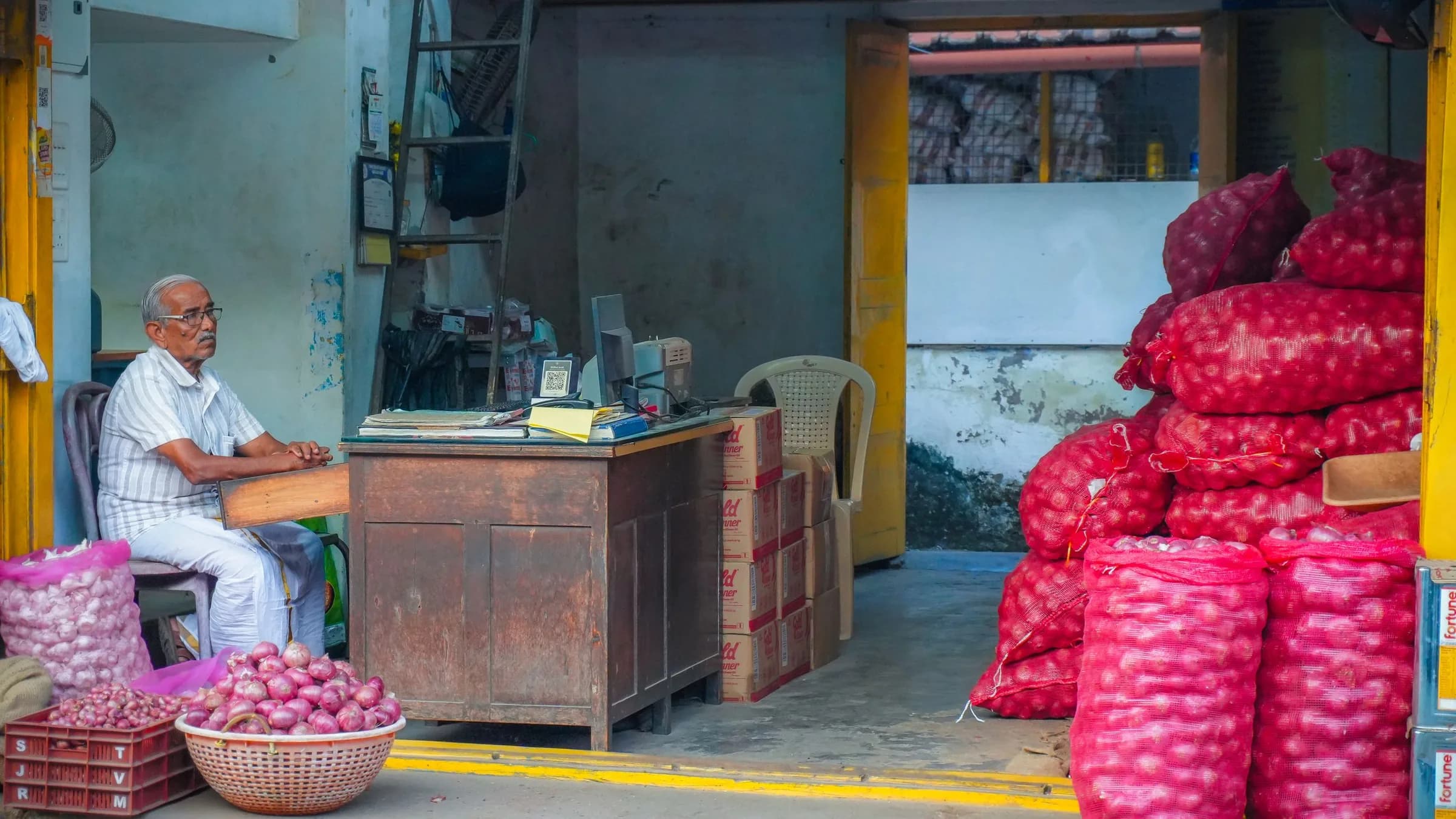 A shop owner sits at a wooden desk at the front of a small produce store, with sacks of red onions stacked high beside him and cartons by the counter.