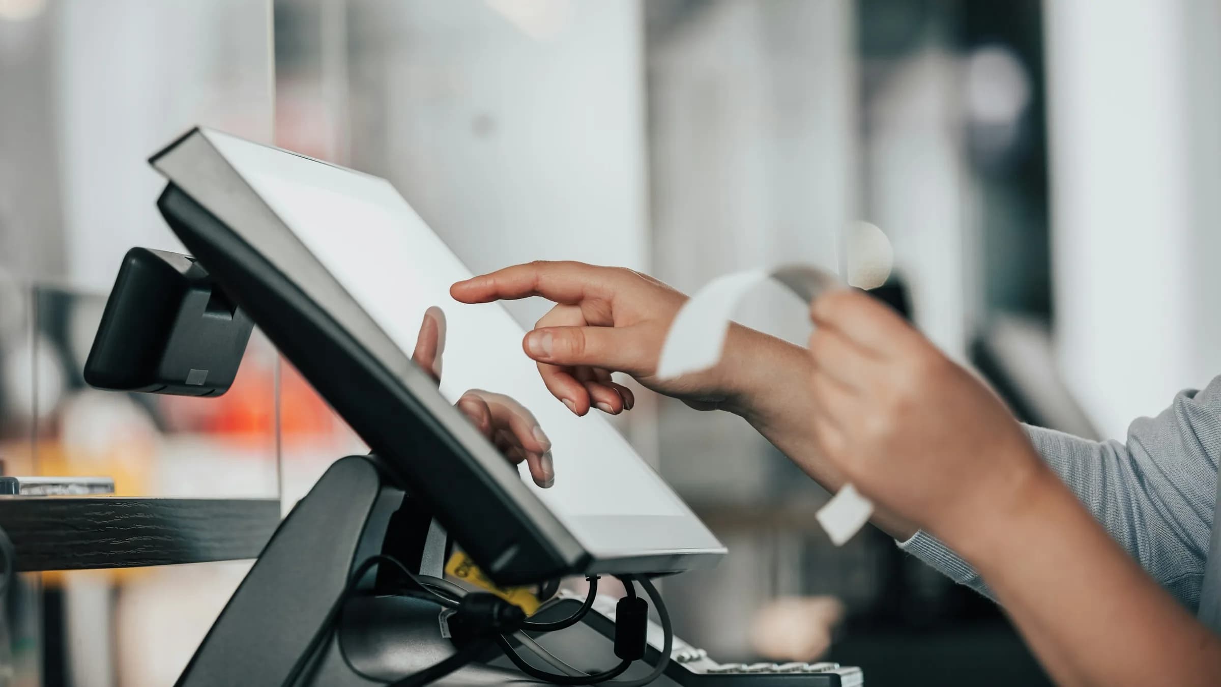 A finger taps the touchscreen of a till on a counter stand while the other hand holds a printed receipt.