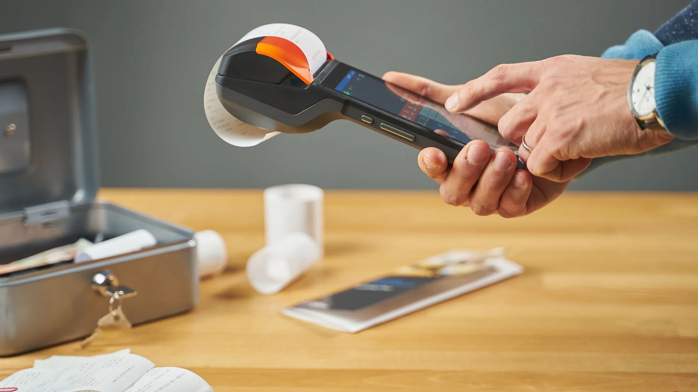 Hands print a receipt from a handheld card terminal above a wooden table with a cash box and spare paper rolls.